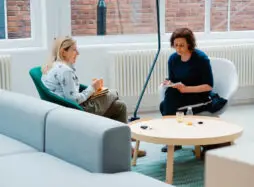 Two women sit facing each other in comfortable chairs, one woman speaks and holds a mug and the other woman takes notes.